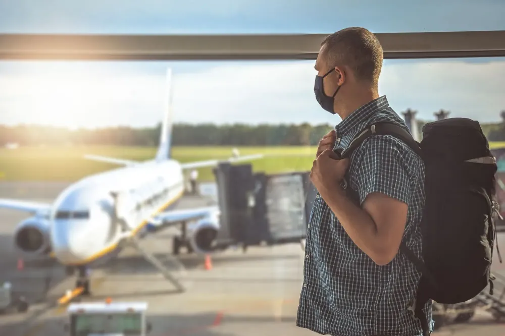 Man wearing a face mask and looking out an airport window at a plane on the tarmac